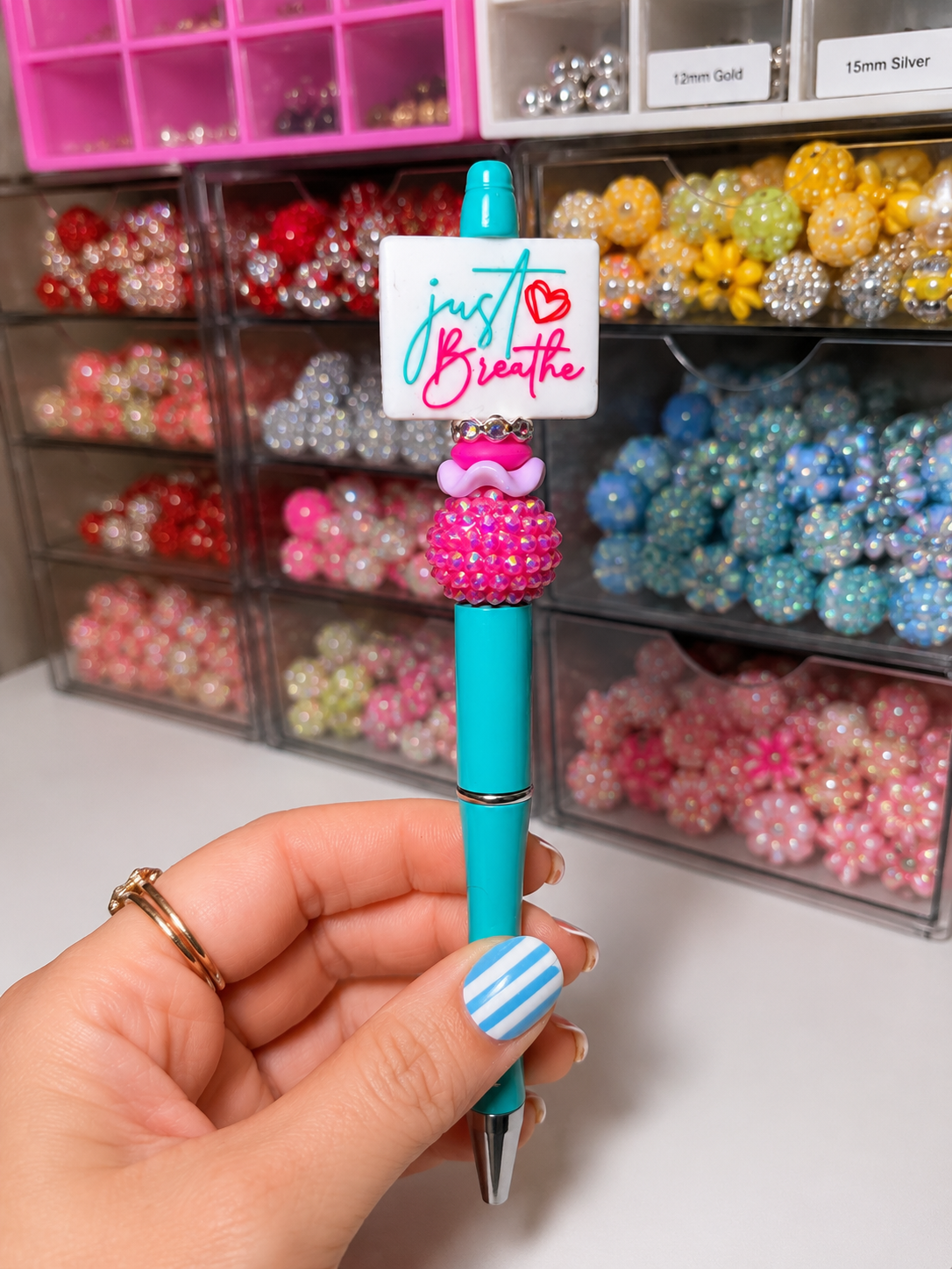 A turquoise ballpoint pen held by a hand, decorated with a white square charm that reads 'just Breathe' in pink and blue script, a pink rhinestone cupcake topper, and a silver clip. The background shows a white desk with clear organizer bins filled with colorful beads.