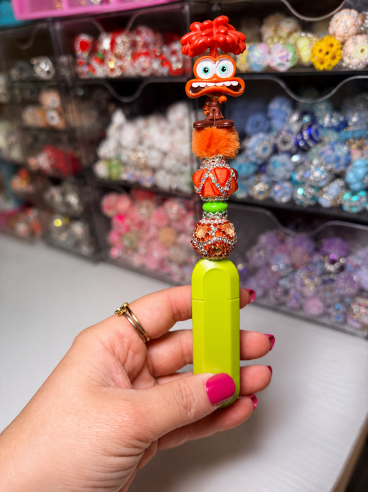 A hand with pink nail polish holds a bright lime green rectangular case decorated with a colorful beaded top featuring a red hair accessory, orange pom-pom, and a cartoon face charm. The background shows shelves filled with colorful jewelry organizer bins.