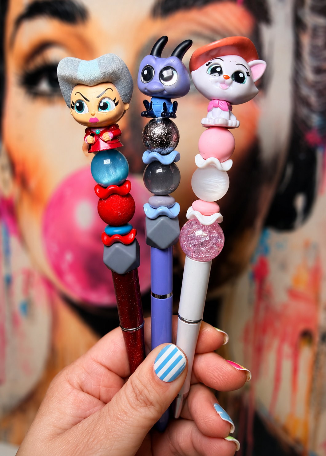 Three decorative novelty pens held in a hand, each topped with a cartoon character figure and colorful bead-like accents, against a blurred graffiti-style background.
