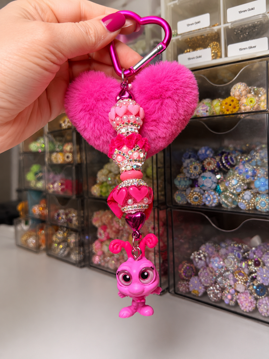 A hand holds a bright pink keychain charm featuring a fluffy heart, a rhinestone-embellished bead, a small pink heart, and a dangling pink character charm, with a background of craft storage bins filled with beads.