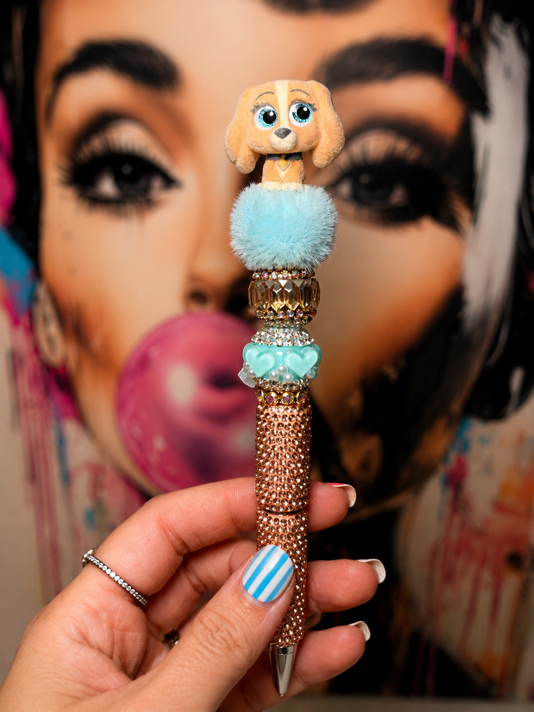 A close-up of a hand holding a decorative pen with a dog character topper, a fluffy light blue pom-pom, rhinestone accents, and a beaded metallic barrel, set against a blurred portrait background.