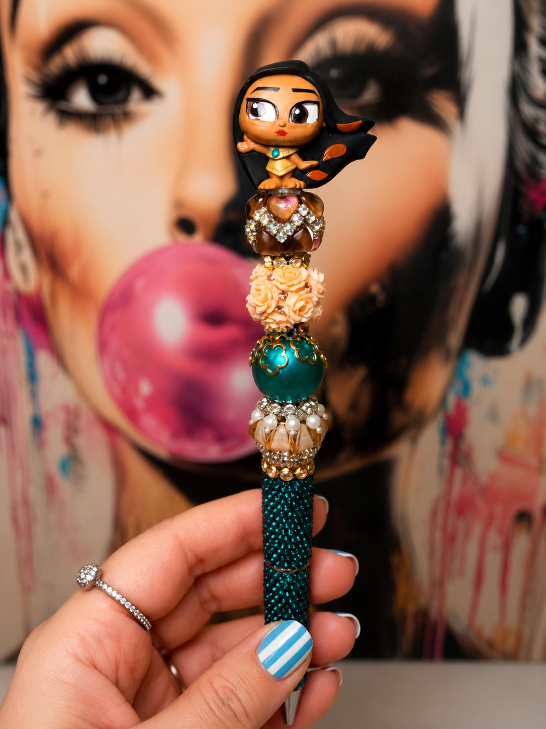A hand holds a decorative beaded pen or stick accessory in front of a colorful art print of a woman blowing pink bubblegum. The product features a small cartoon character at the top, rhinestone details, peach and teal beads, and a dark teal beaded lower section.