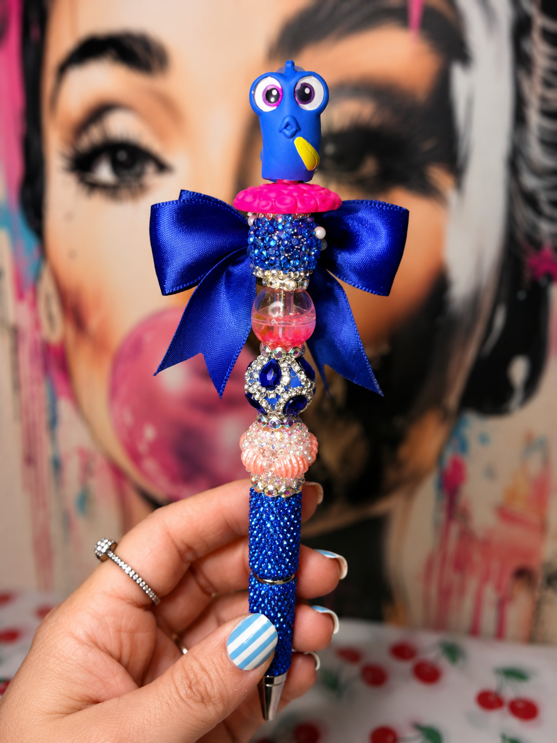 A hand holds a decorative blue pen featuring a cartoon bird figure, a large blue satin bow, and embellished beads, with a colorful painted face in the background.