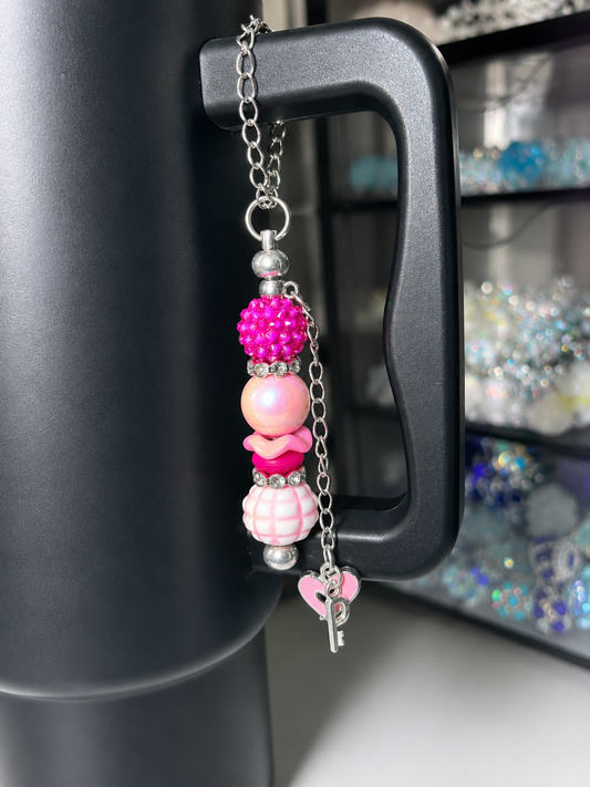 A pink and silver beaded keychain charm with a silver key clasp, hanging from the black handle of a dark gray mug, with a blurred craft storage organizer in the background.