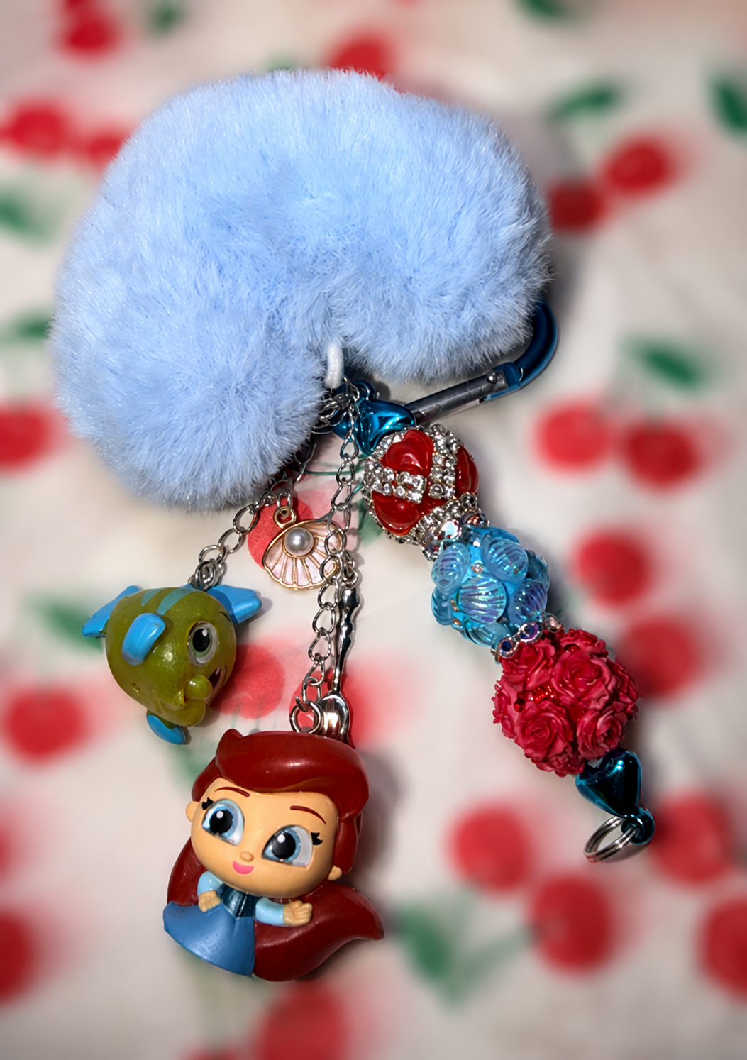 A light blue fluffy pom-pom keychain with a carabiner clip and multiple dangling charms, including a red-haired character figure, a green fish, and colorful bead balls, shown against a blurred red-and-white patterned background.