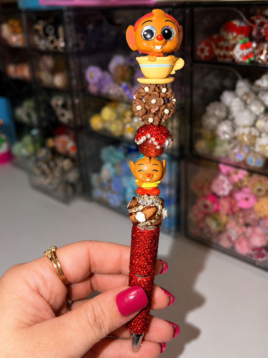 A hand with pink nail polish holds a sparkly red pen decorated with two cartoon owl characters in gold and orange, stacked on top of brown beads and a red glitter ball, against a blurred background of craft supplies.