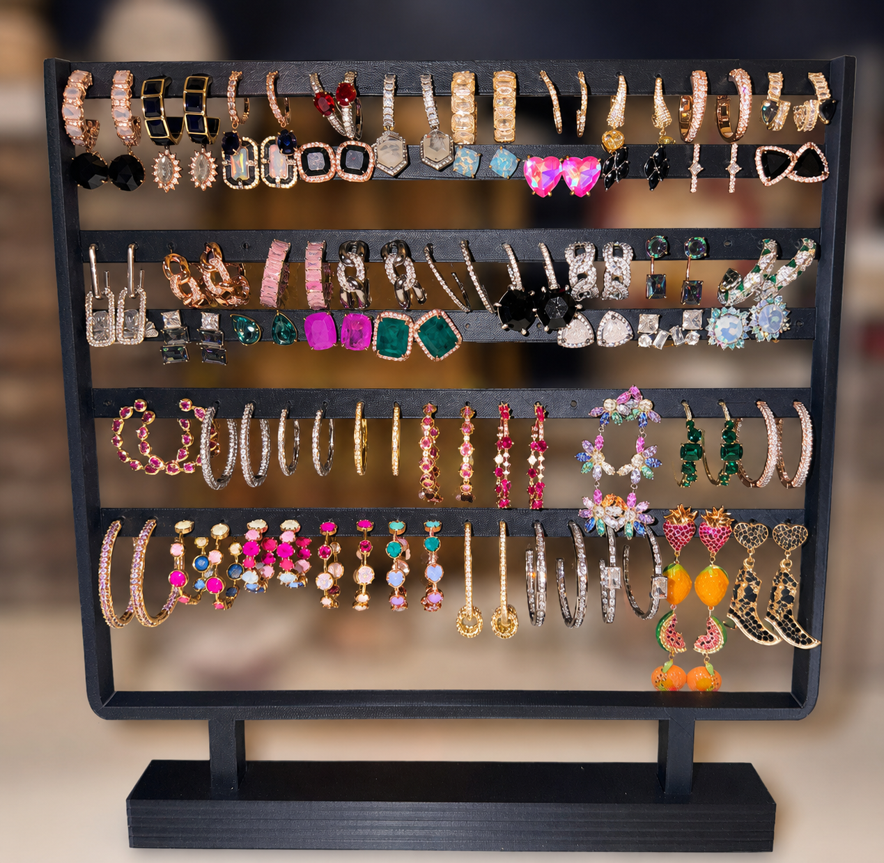 A black metal multi-tier jewelry display stand filled with assorted earrings and bracelets, photographed against a softly blurred warm indoor background.