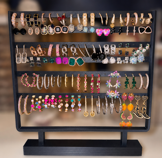 A black metal multi-tier jewelry display stand filled with assorted earrings and bracelets, photographed against a softly blurred warm indoor background.