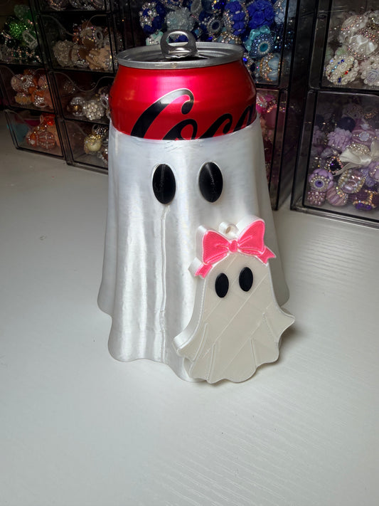 A Halloween decoration shaped like a white ghost draped over a red Coca-Cola can, with black eyes and a small pink bow on a white ornament in front, set on a white surface with storage drawers in the background.