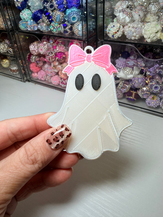 A hand with leopard-print nail polish holds a 3D-printed ghost charm with a pink bow, with a background of clear organizer drawers filled with craft supplies.