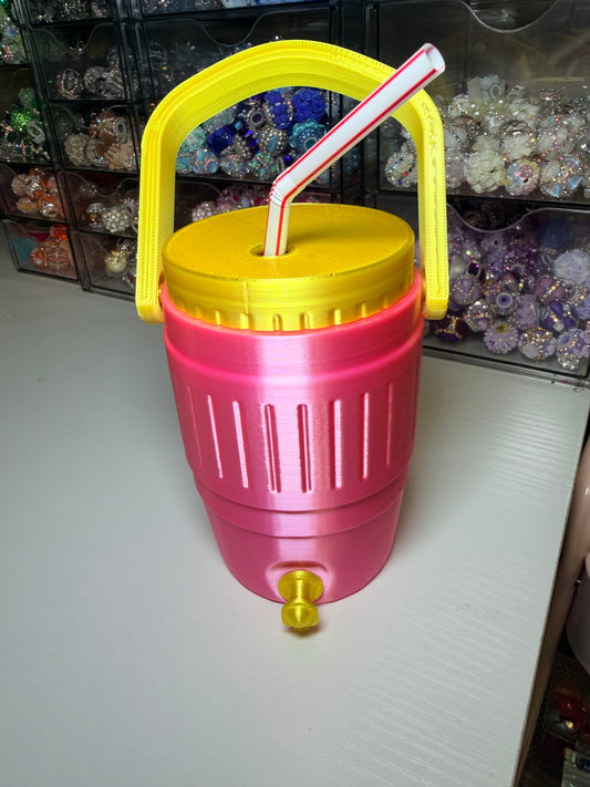 A novelty pink and yellow plastic beverage dispenser with a yellow handle, yellow lid, and a red-and-white striped straw, sitting on a white tabletop with storage bins in the background.
