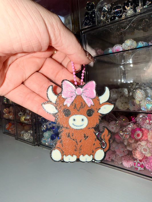 A hand holds a small brown cow-shaped charm or keychain with white horns and feet, a pink bow, and a pink beaded loop, in front of shelves filled with colorful craft supplies.