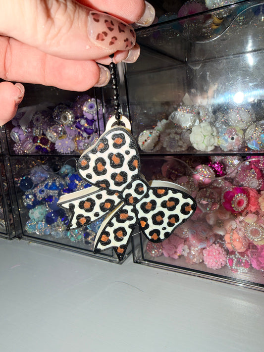 A hand holds a small bow-shaped accessory with a white, black, and orange leopard print pattern against a background of clear plastic drawers filled with colorful craft embellishments.