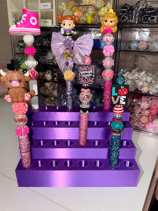 A purple 3D-printed tiered display stand holding multiple decorative beaded pens or nail art sticks with colorful charms, shown on a white desk with storage drawers in the background.