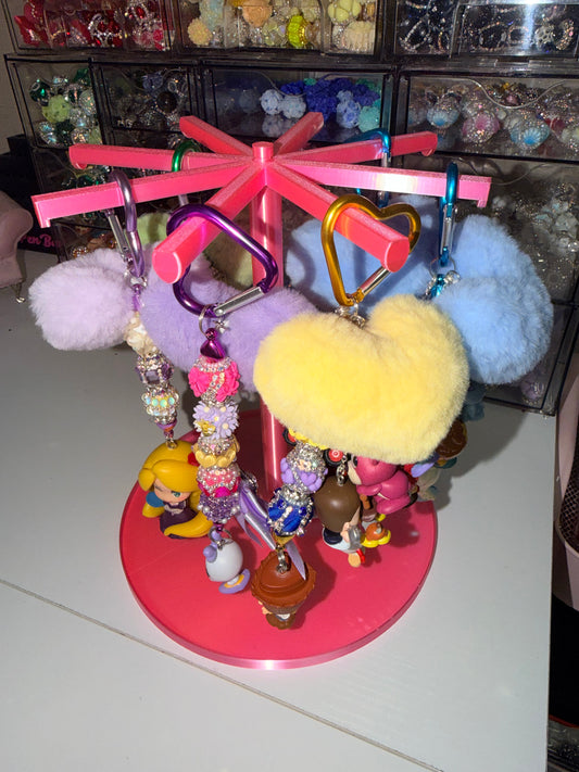 A pink rotating keychain display stand filled with colorful charms, pom-poms, and small figurines, set on a white tabletop with storage bins in the background.