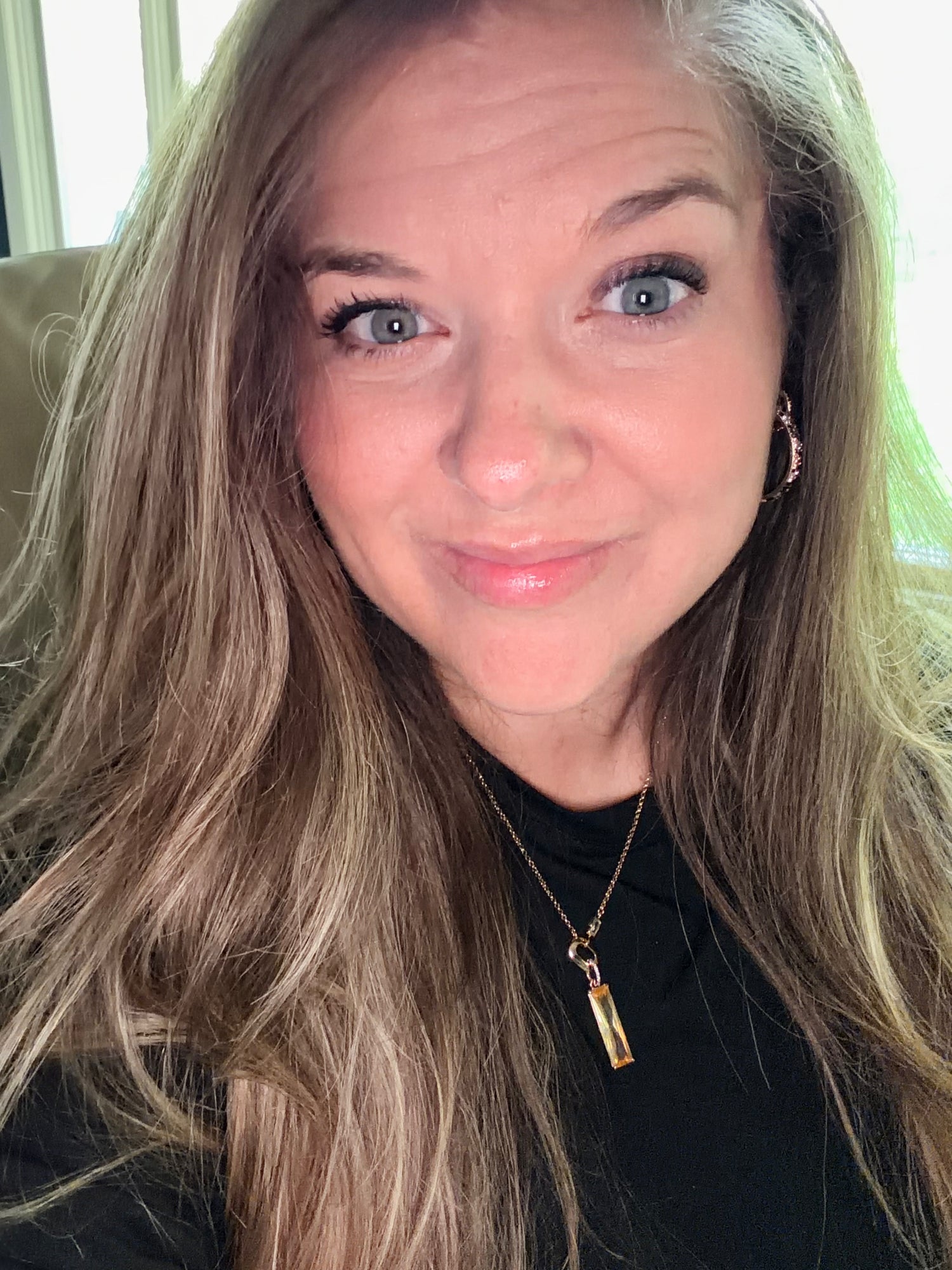 A close-up portrait of a woman with long blonde hair, wearing a black top and a gold pendant necklace, photographed indoors.