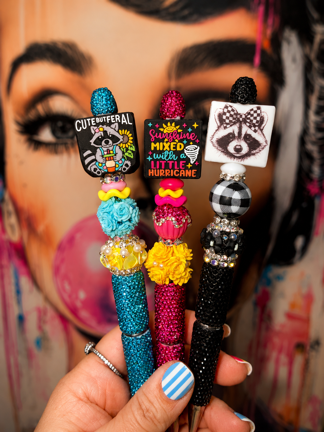 Three decorative pen-style accessories held in a hand, featuring colorful glitter handles, floral embellishments, rhinestone accents, and square charms with raccoon illustrations and text. The background shows a painted portrait with bold colors.