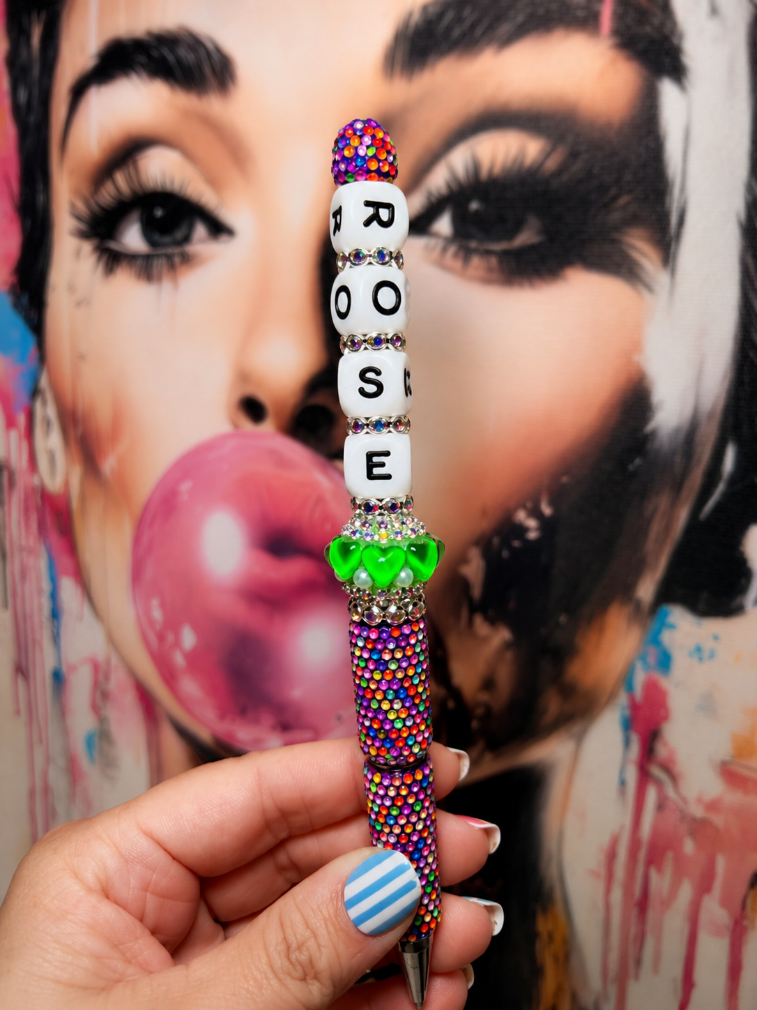 A hand holds a custom beaded pen in front of a colorful pop-art portrait of a woman blowing a pink bubblegum bubble. The pen features white letter beads spelling 'ROOSE' with rhinestone accents, a green heart-shaped charm, and a multicolor beaded barrel.