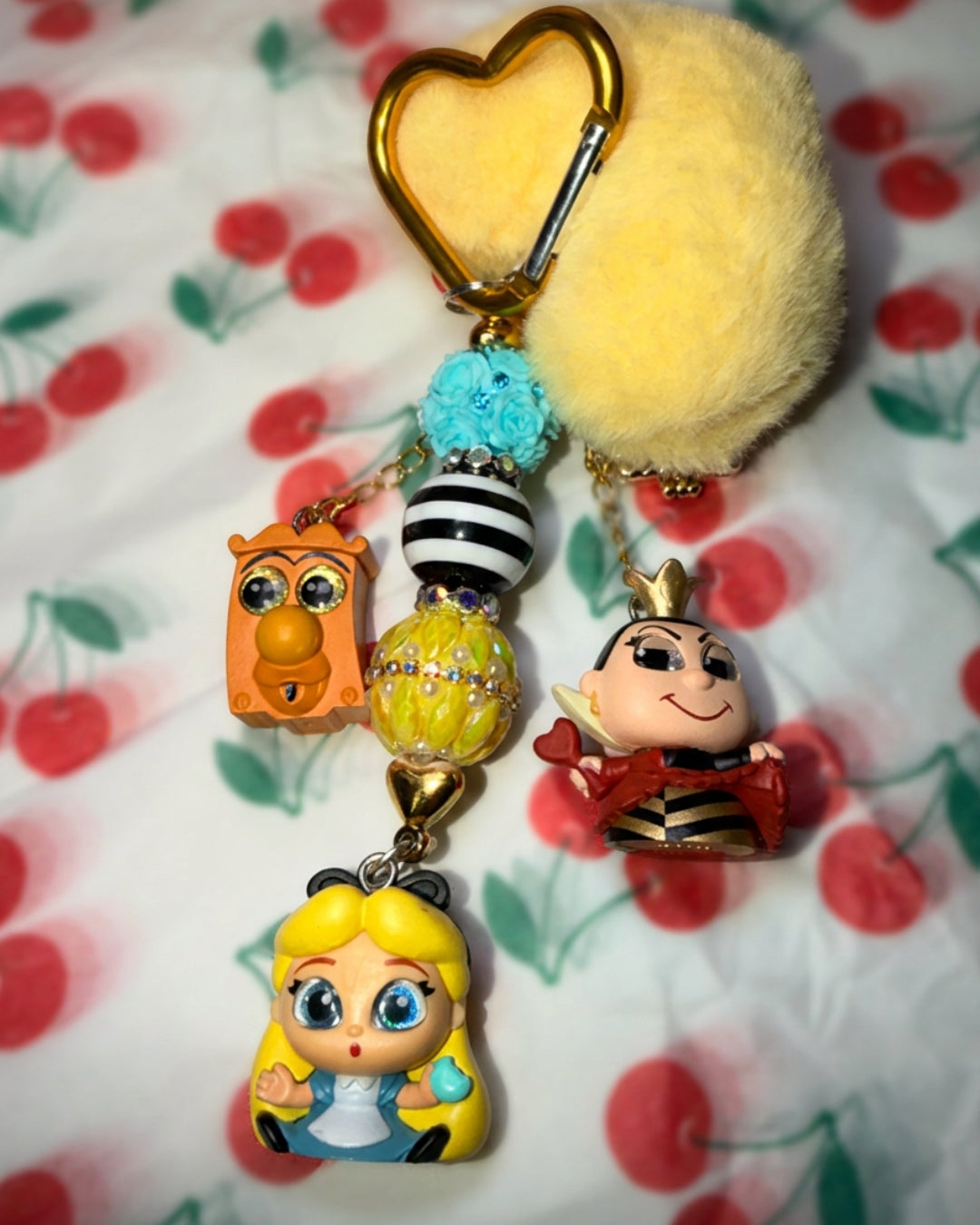 A colorful keychain charm featuring a gold heart-shaped clasp, a fluffy yellow pom-pom, a blue bead, and several character charms including Alice, the Queen of Hearts, and an owl figure, displayed on a cherry-patterned fabric background.